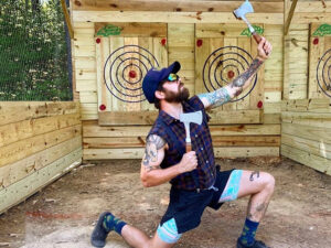 A man posing triumphantly with throwing axes in front of a target in Williamsburg, Virginia